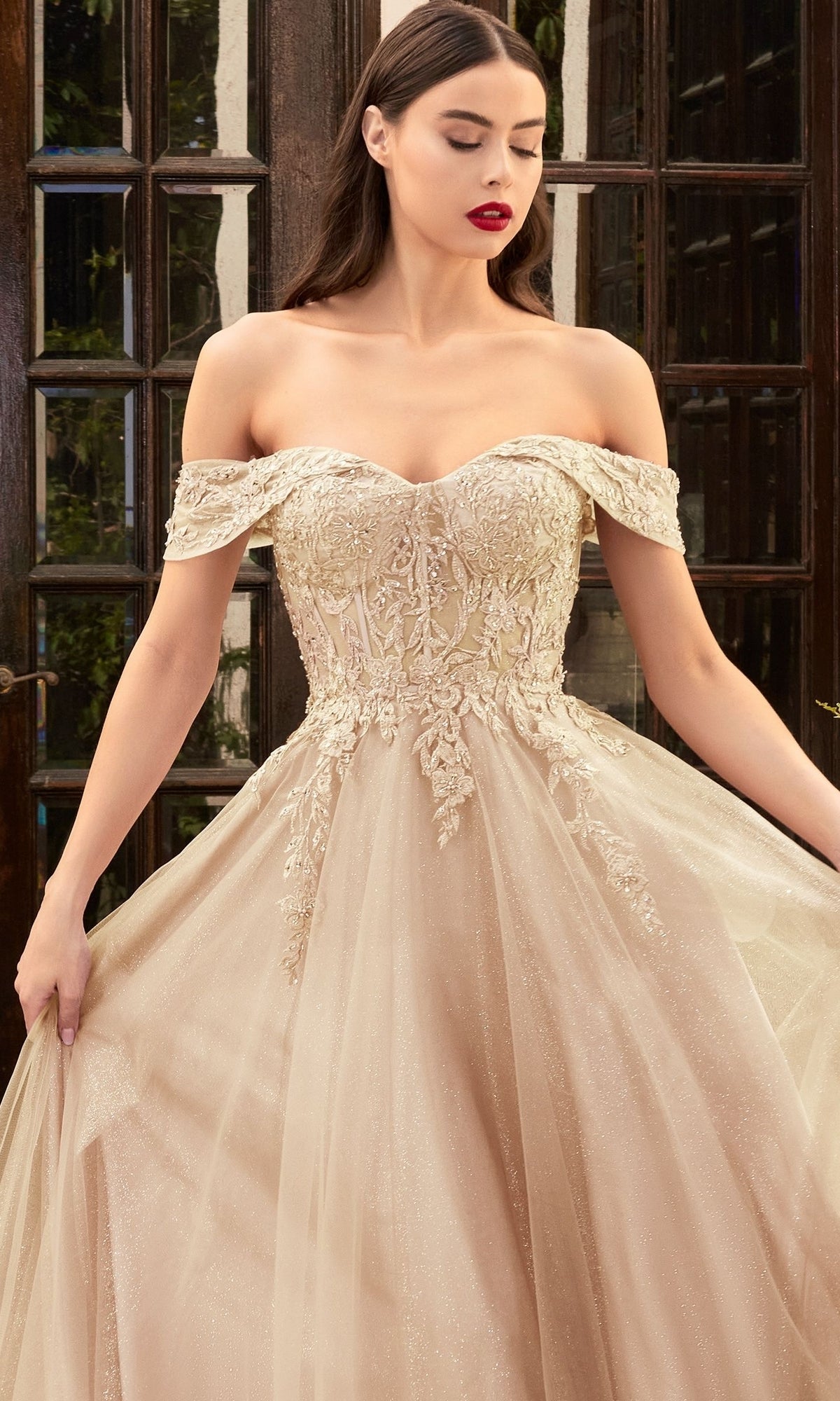 A woman wearing the Sheer-Bodice Long Glitter Prom Ball Gown CD961, with elegant embroidery and a tulle skirt, stands in front of glass doors.